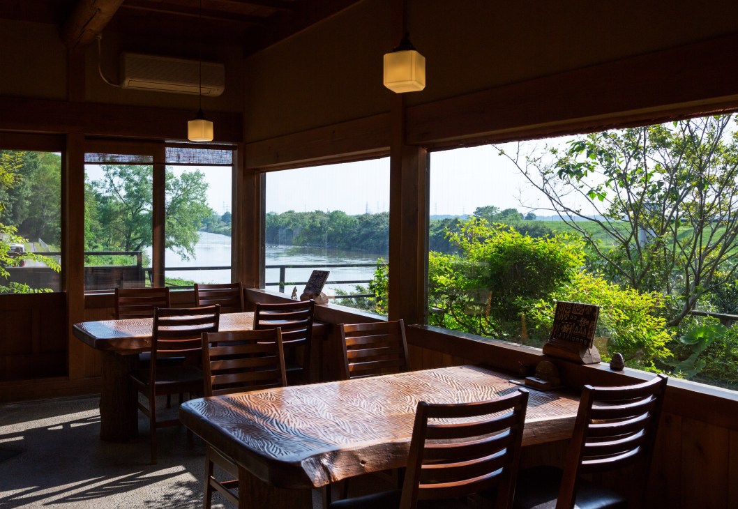 Dining room with wooden tables and chairs overlooking a river through large windows.