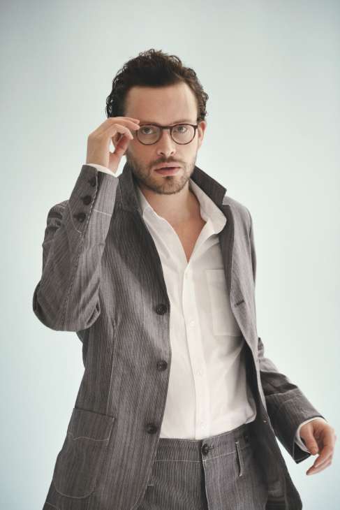 Man wearing grey pinstriped suit jacket and white shirt with round-framed glasses.