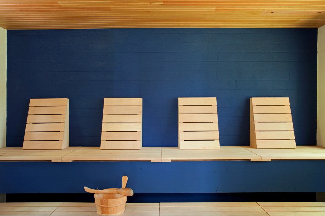 Sauna interior with four wooden backrest benches against a deep blue wall and wooden bucket with ladle.