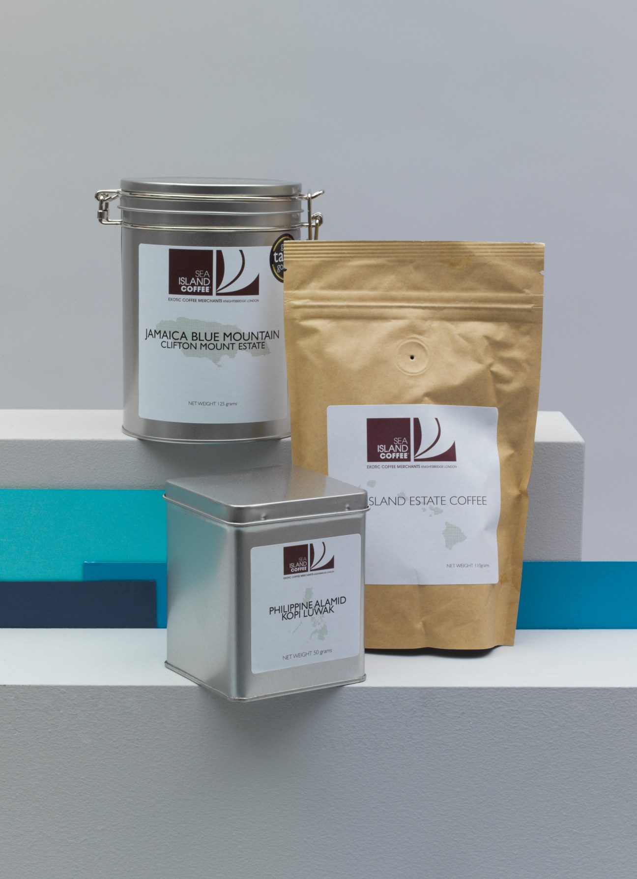 Sea Island Coffee packaging displayed in three containers: a metal tin with handle, a smaller metal tin, and a kraft paper pouch.