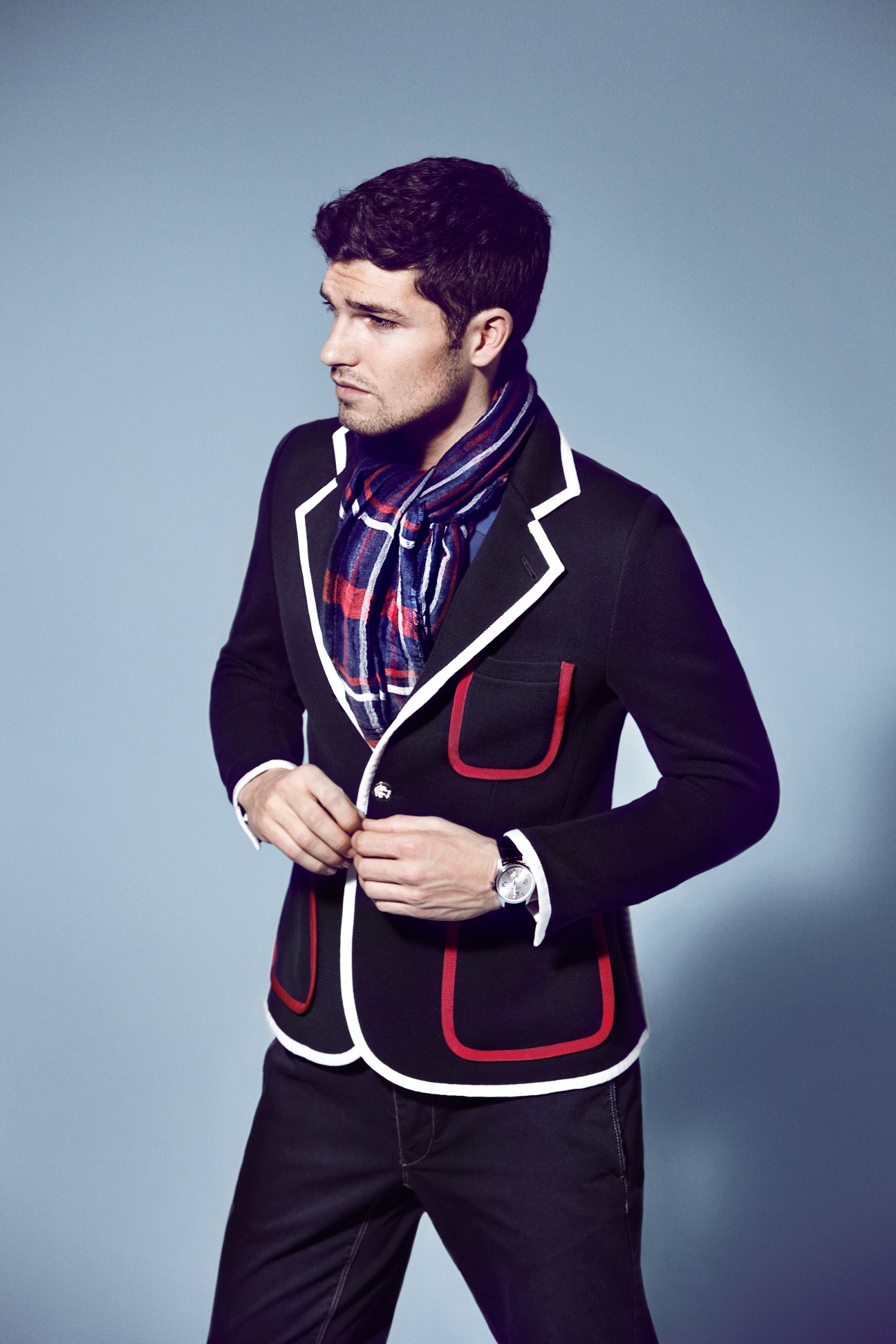 Man wearing Black Fleece by Brooks Brothers jacket with red and white trim, Lacoste polo shirt, and Omega watch against blue background.