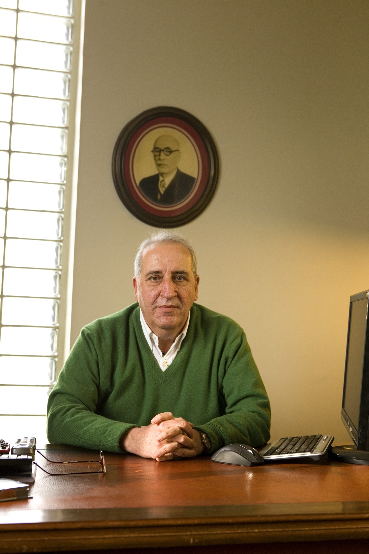 Ali Muhsinzade sits at a desk wearing a green sweater, with a portrait hanging on the wall behind him.