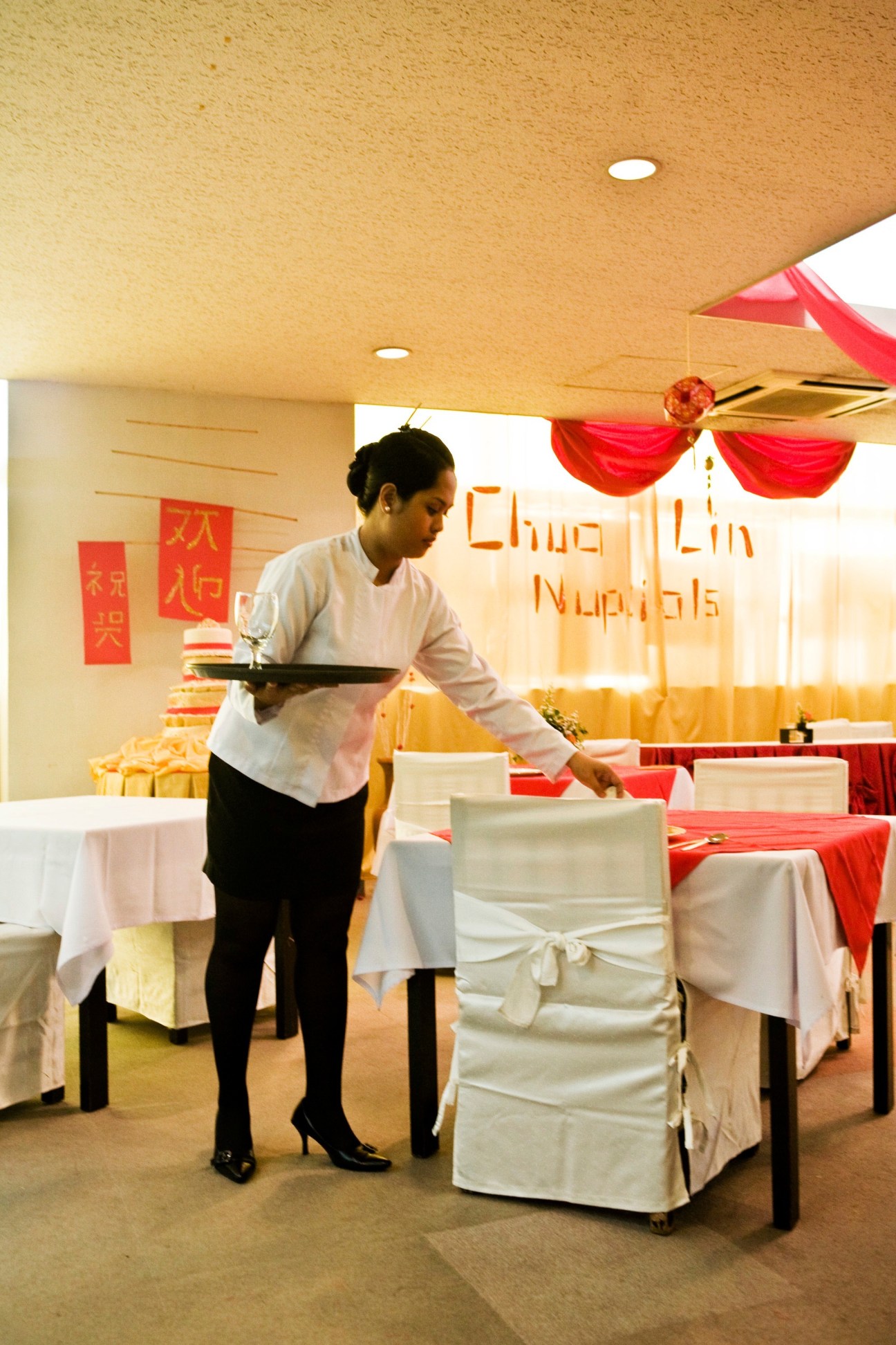A hospitality student practices serving skills in a mock dining room with red and white table settings and Chinese decorations.