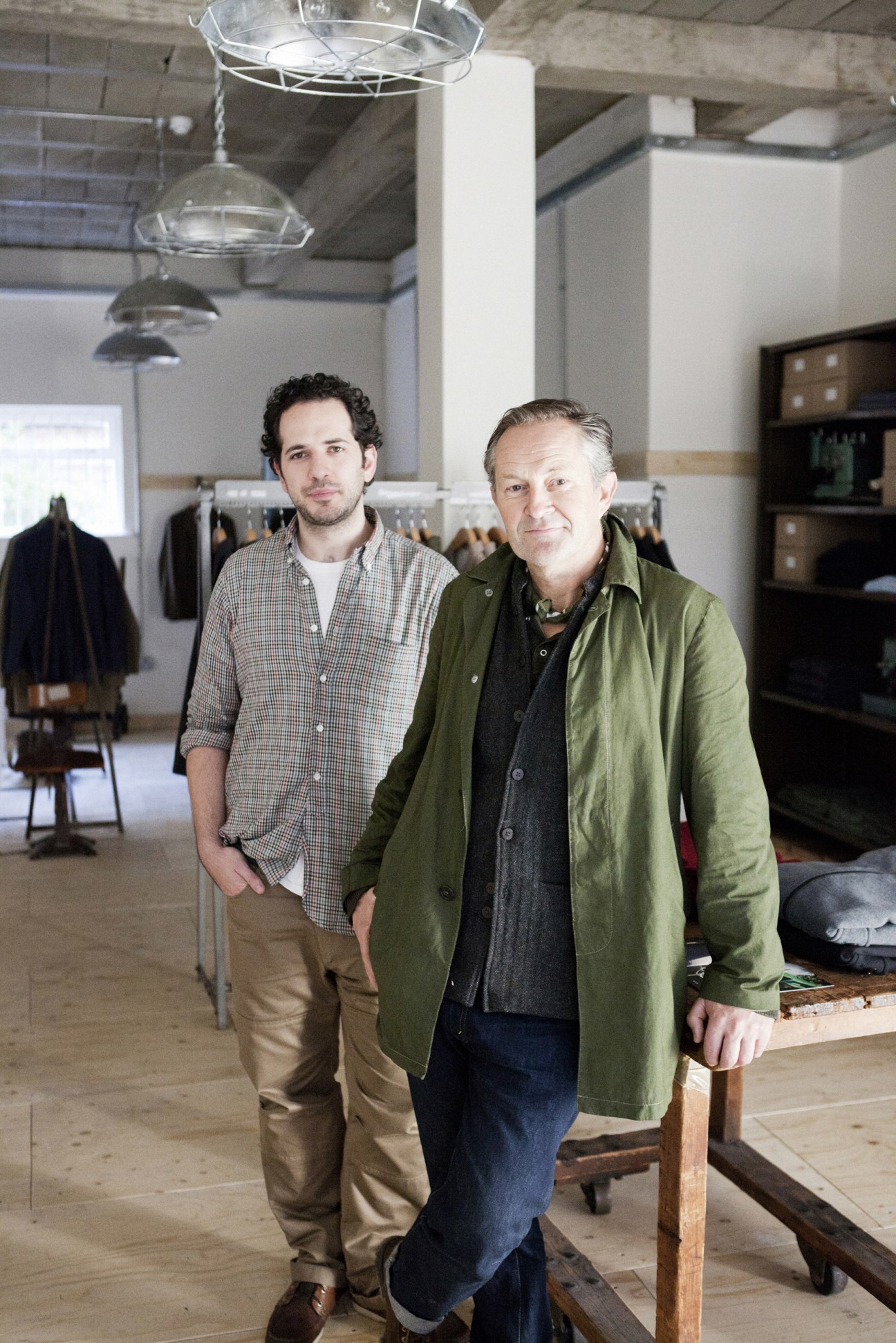 Two men stand in a minimalist clothing store with pendant lighting and wooden shelves.