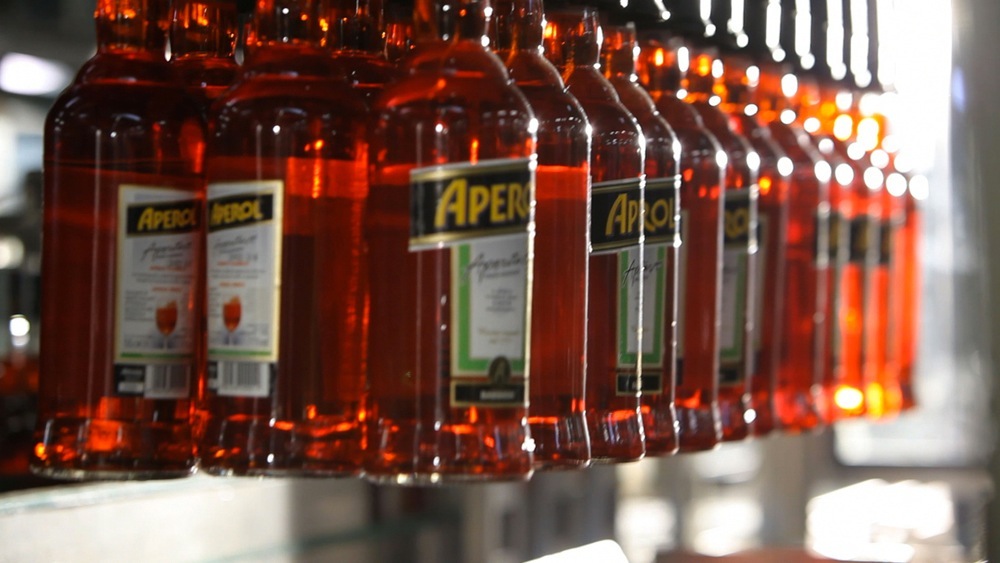 Row of Aperol liqueur bottles with amber-colored liquid and yellow labels on a production line.
