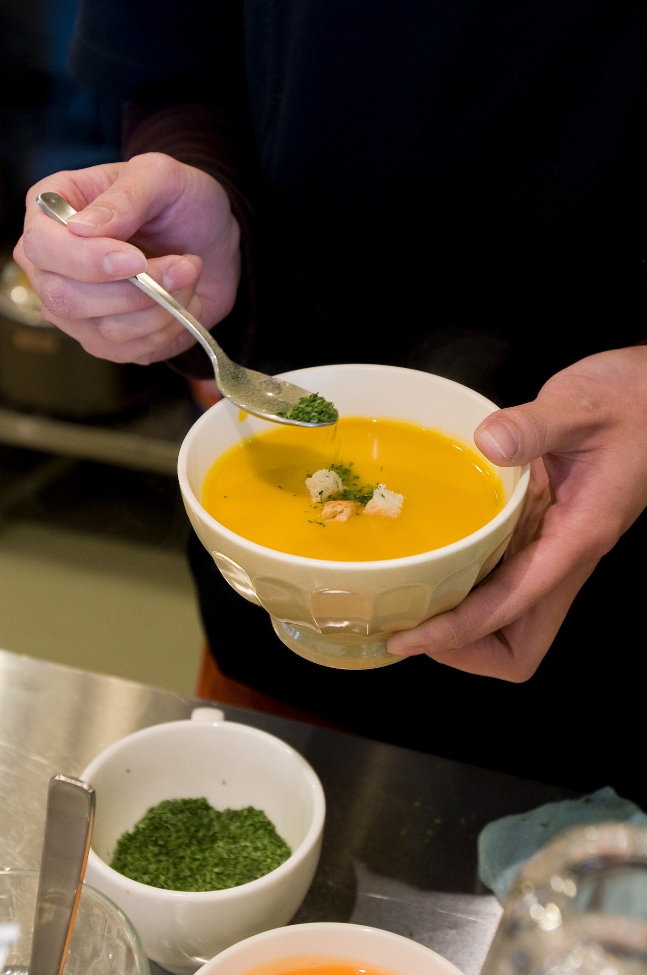 Hands holding a white bowl of golden soup garnished with green microgreens and white protein, with a spoon mid-dip.