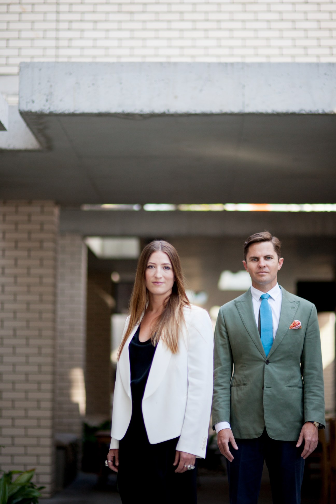 Two architects, a woman in a white blazer and a man in a green jacket, stand in front of a modern concrete building.