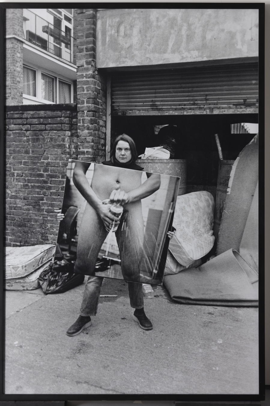 Black and white photograph of a person holding an explicit artwork outside a brick building.