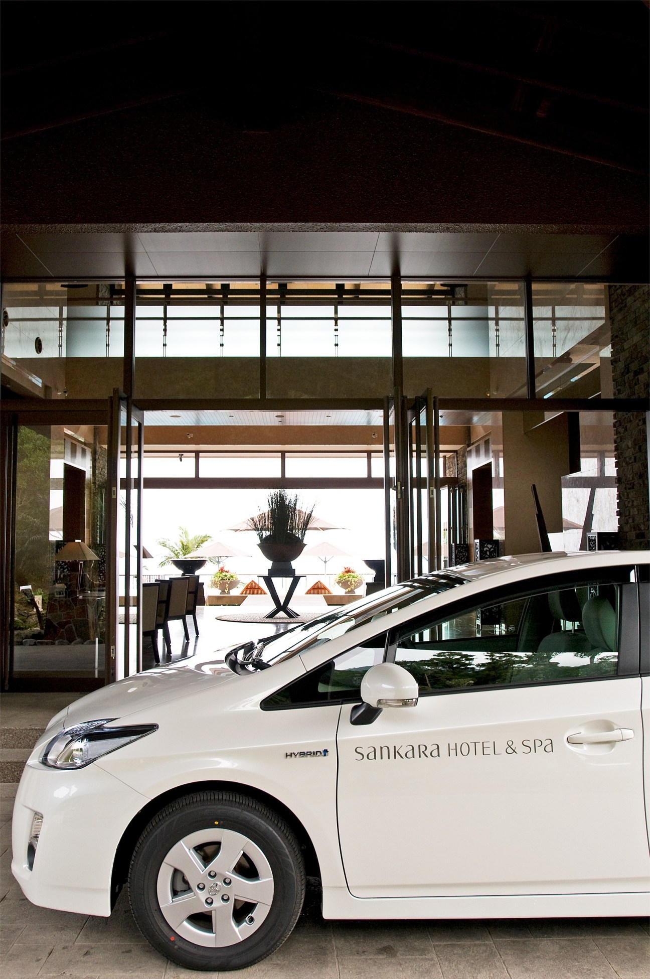 White hybrid Prius car parked outside Sankara Hotel & Spa entrance with modern glass doors and terrace visible.
