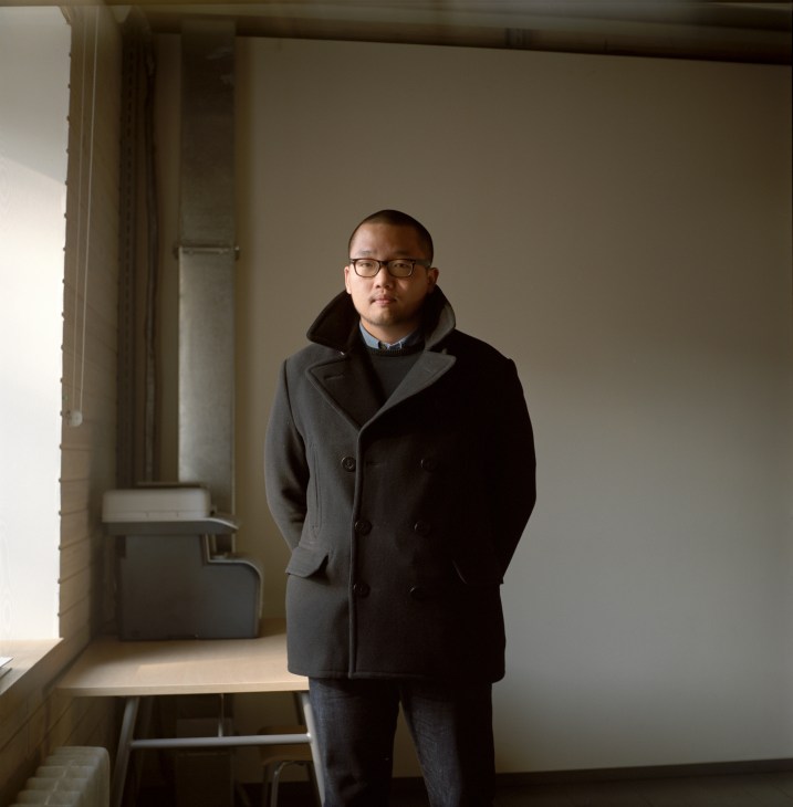 Strelka student Kang Minkoo standing in a minimalist interior space wearing a dark coat and glasses.