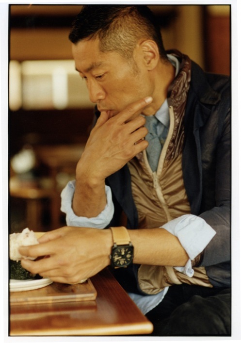 Man wearing Sacai layered jacket and Engineered Garments shirt touching his face while seated indoors.