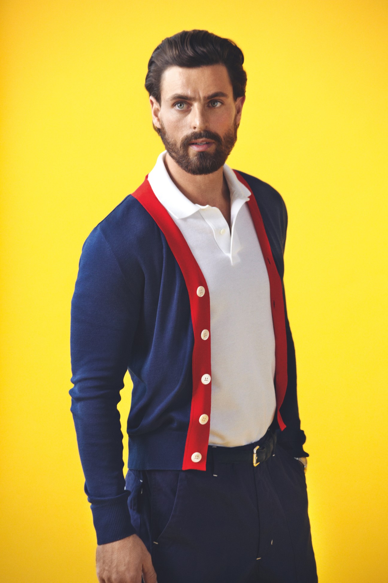 Man wearing a navy cardigan with red stripe, white polo shirt, and black trousers against yellow background.