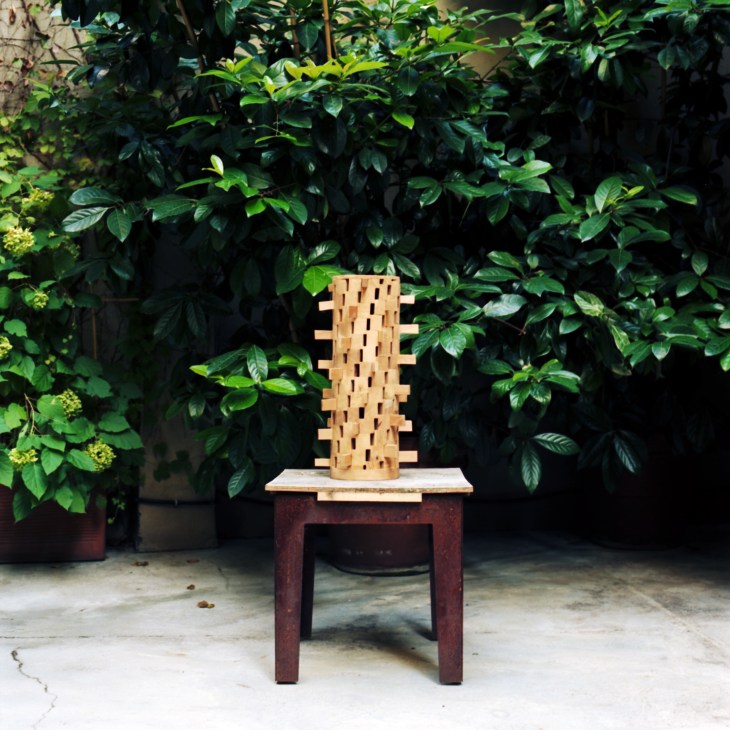 Wooden architectural model of a tower with perforated sides displayed on a red stool in a garden.