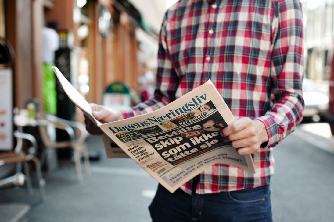 Person in red plaid shirt reading Dagens Næringsliv newspaper on a street