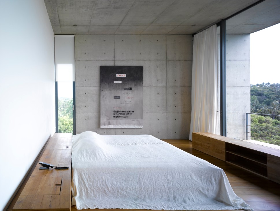 Minimalist bedroom with white bed, bare concrete walls, and large windows overlooking jungle landscape.
