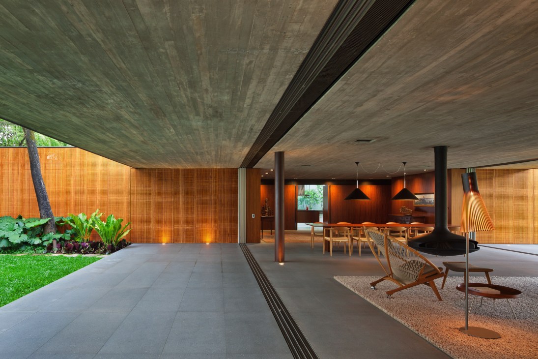 Modern open-plan living room with retractable glass walls opening to garden, featuring mid-century furniture and suspended fireplace.