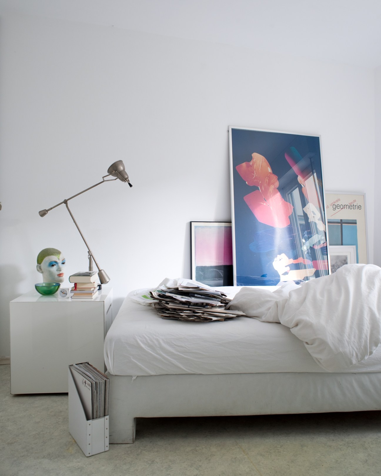 A minimalist bedroom with white bedding, a desk lamp, colorful artworks, and stacked magazines on the bed.