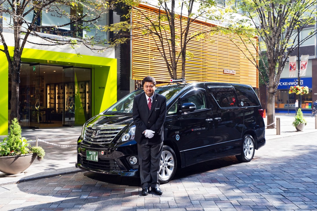 A professional taxi driver in formal attire stands beside a black Toyota Alphard vehicle on a city street.