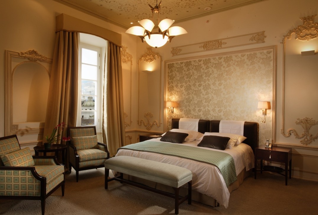 Elegant hotel bedroom with ornate cream and gold decor, featuring a bed with black headboard and green throw, matching armchairs, and a chandelier.
