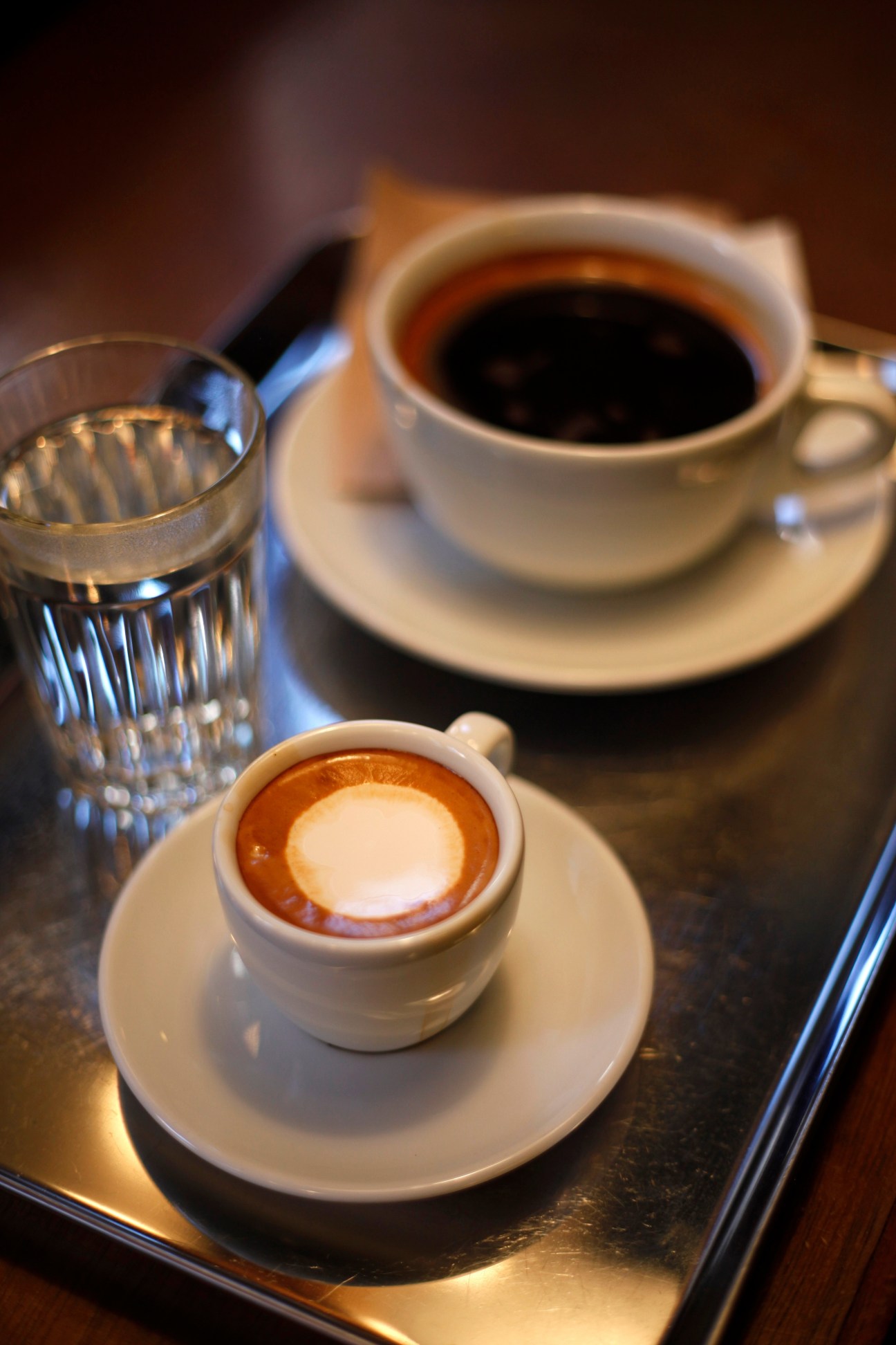 A cappuccino with latte art and a glass of water on a dark table, with a black coffee in the background.