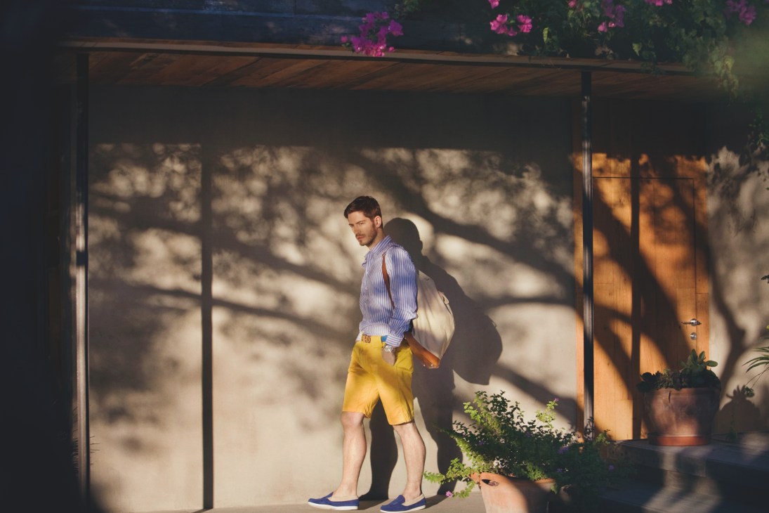 Man in striped shirt and yellow shorts standing under a pergola with purple bougainvillea at Macakizi hotel in Bodrum.