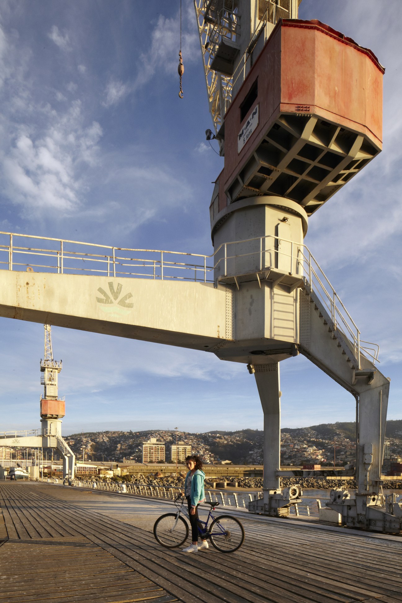 Cyclist with bike standing on wooden pier beneath industrial cargo crane at Valparaiso's Muelle Barón port.