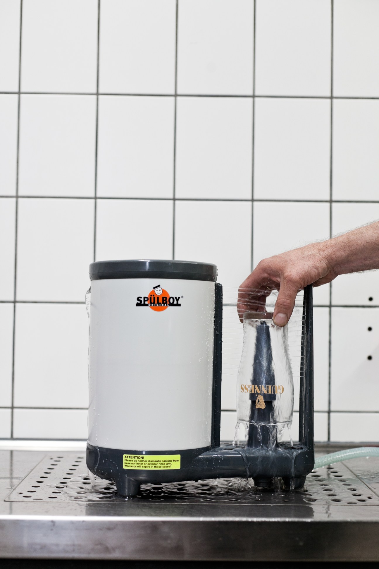 A hand demonstrates the Spülboy glass washer in action, showing a clean beer glass being removed from the white and black machine.