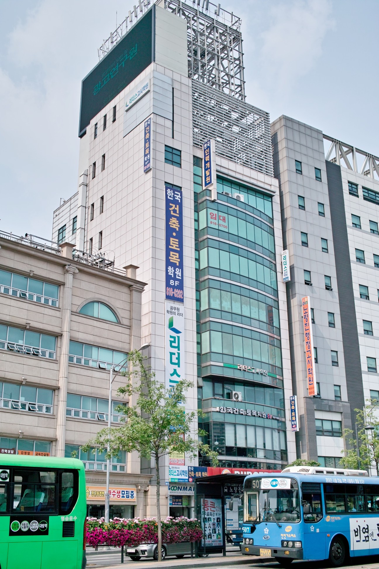 A tall multi-story building in Seoul with numerous Korean-language hagwon signs and advertisements displayed on its facade, with buses and street-level activity visible below.