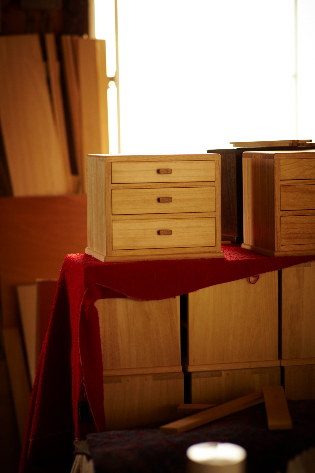 Wooden furniture piece with three drawers displayed on a table with red fabric draping in warm lighting.