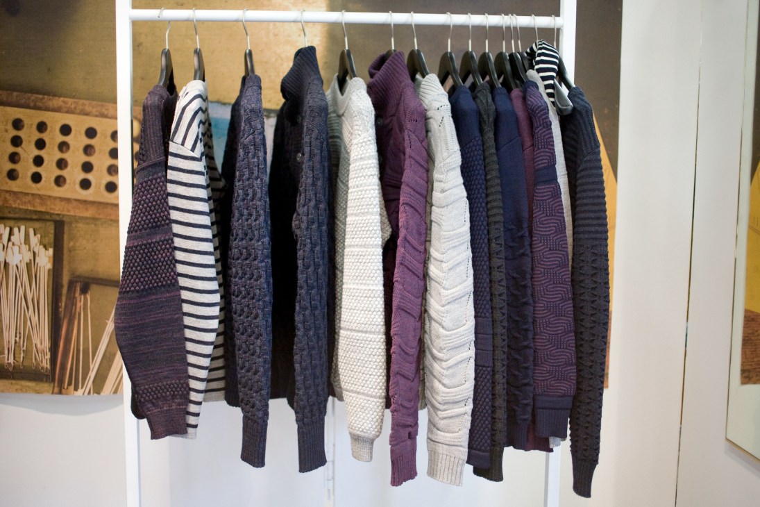 SNS Herning knitwear collection hanging on a white rail, displaying various knitted sweaters in navy, purple, white and striped patterns.