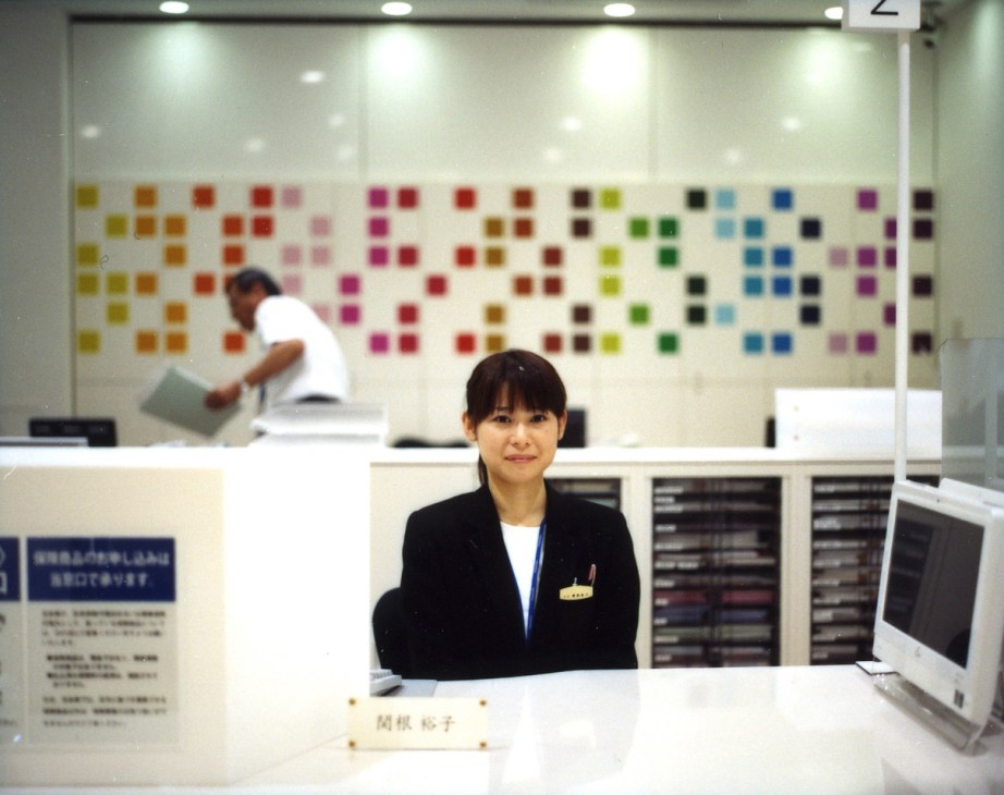 Bank teller Yuko Sekine at her desk with colorful squares displayed on the wall behind her.