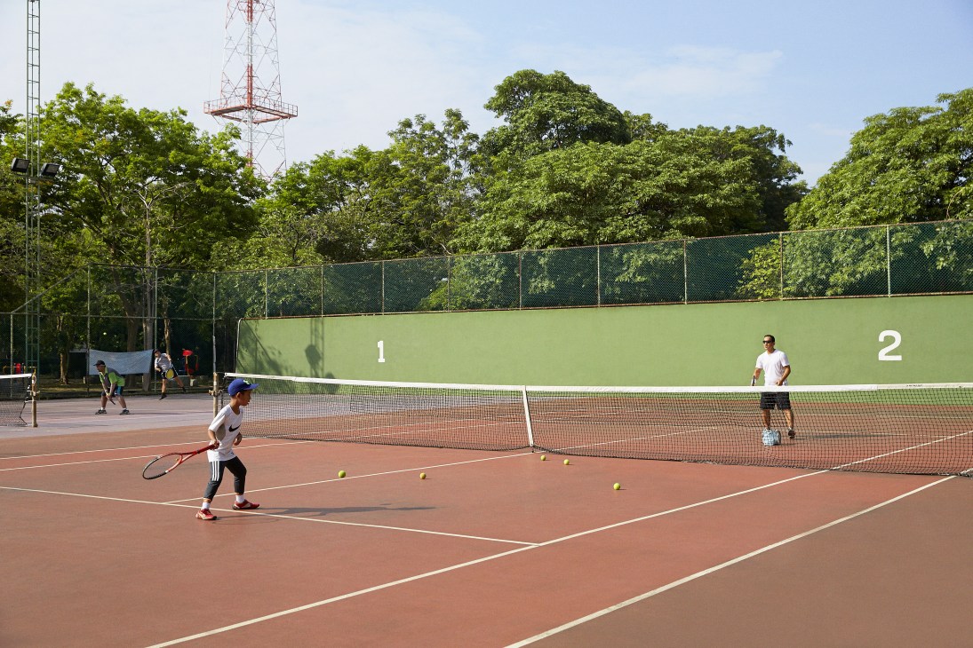 Father and son playing tennis at Ari tennis club in Bangkok with numbered court walls and trees in background.