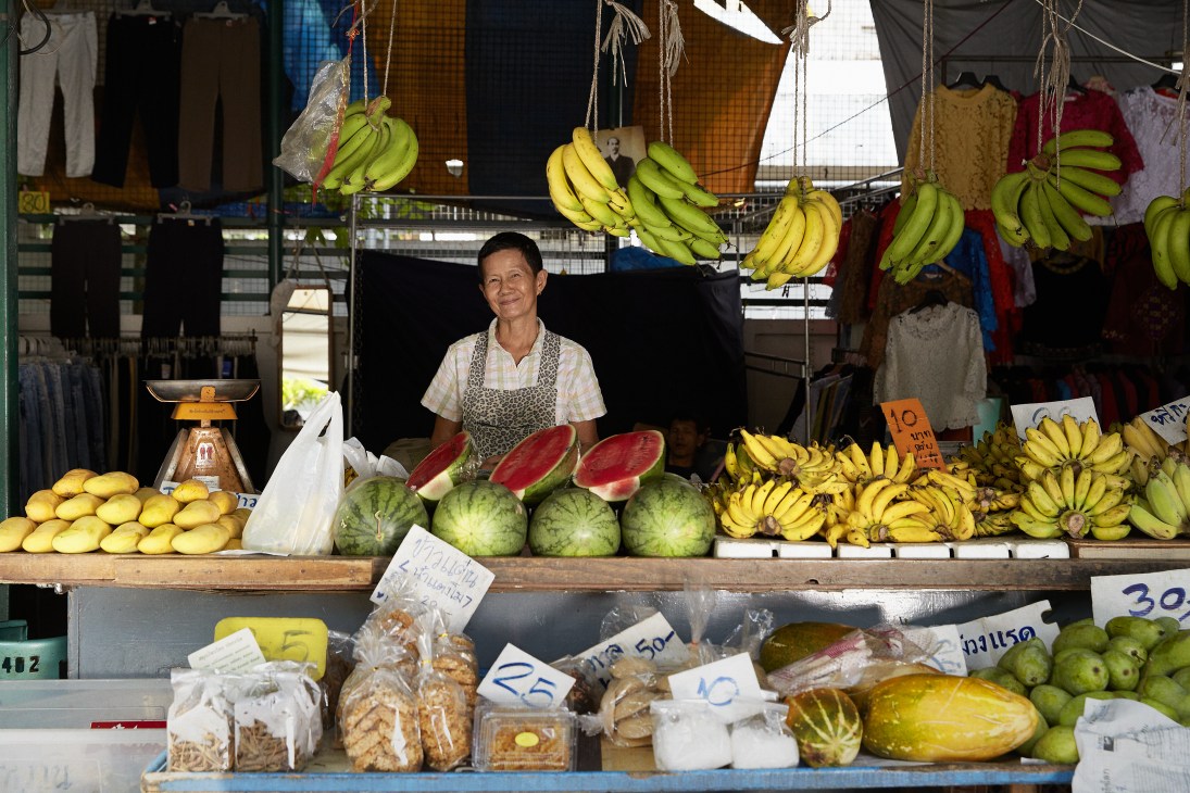 Woman vendor at Bangkok market stall displaying hanging bunches of bananas and tropical fruits