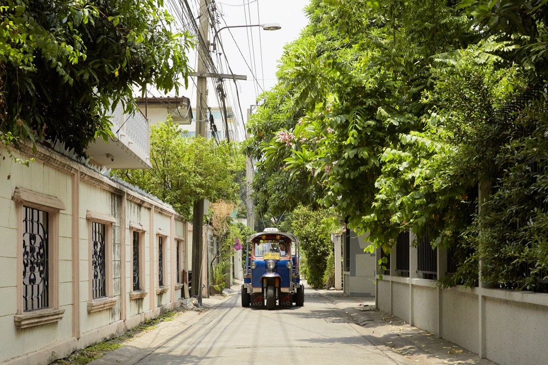 A blue Thai tuk-tuk navigating a narrow, tree-lined street in Bangkok's Ari neighborhood.