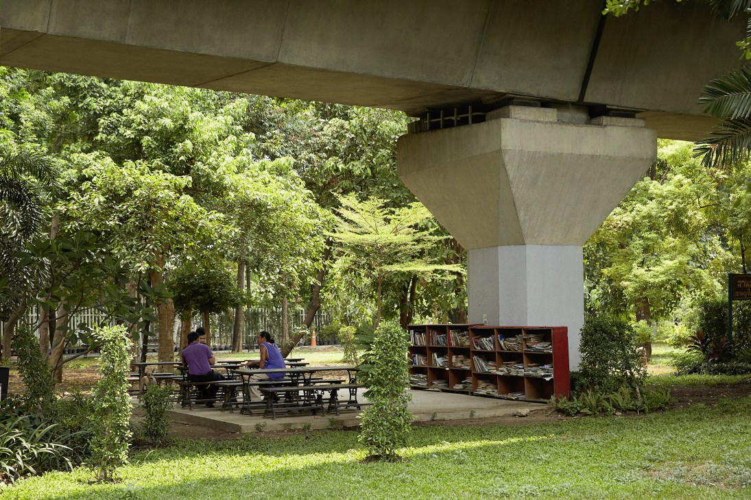 Two people sit at picnic tables beneath a concrete overpass in a lush green park with an open-air library structure.