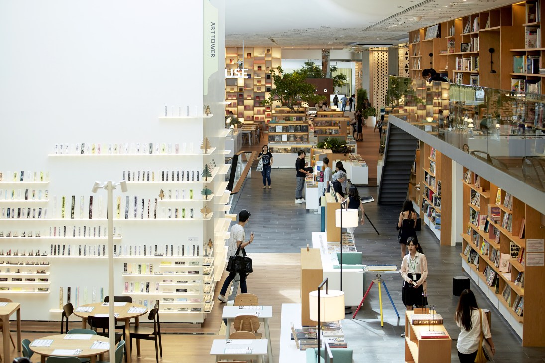 Interior view of Open House showing multi-level wooden shelving with books, plants, and visitors browsing in Bangkok's Central Embassy.