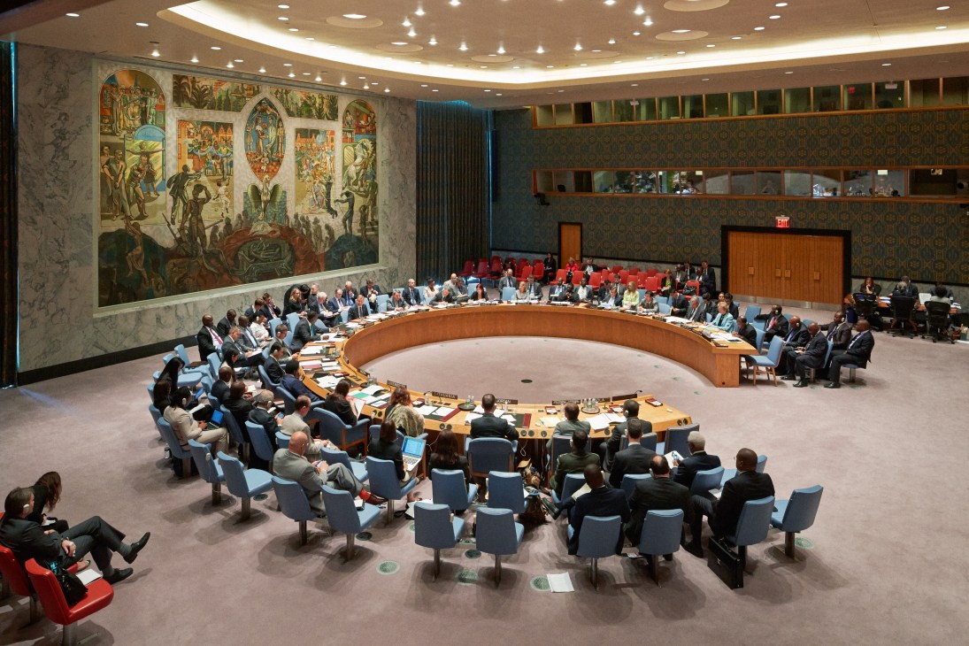 The UN Security Council chamber with diplomats seated around a curved horseshoe table during a session, with interpreter booths visible above.