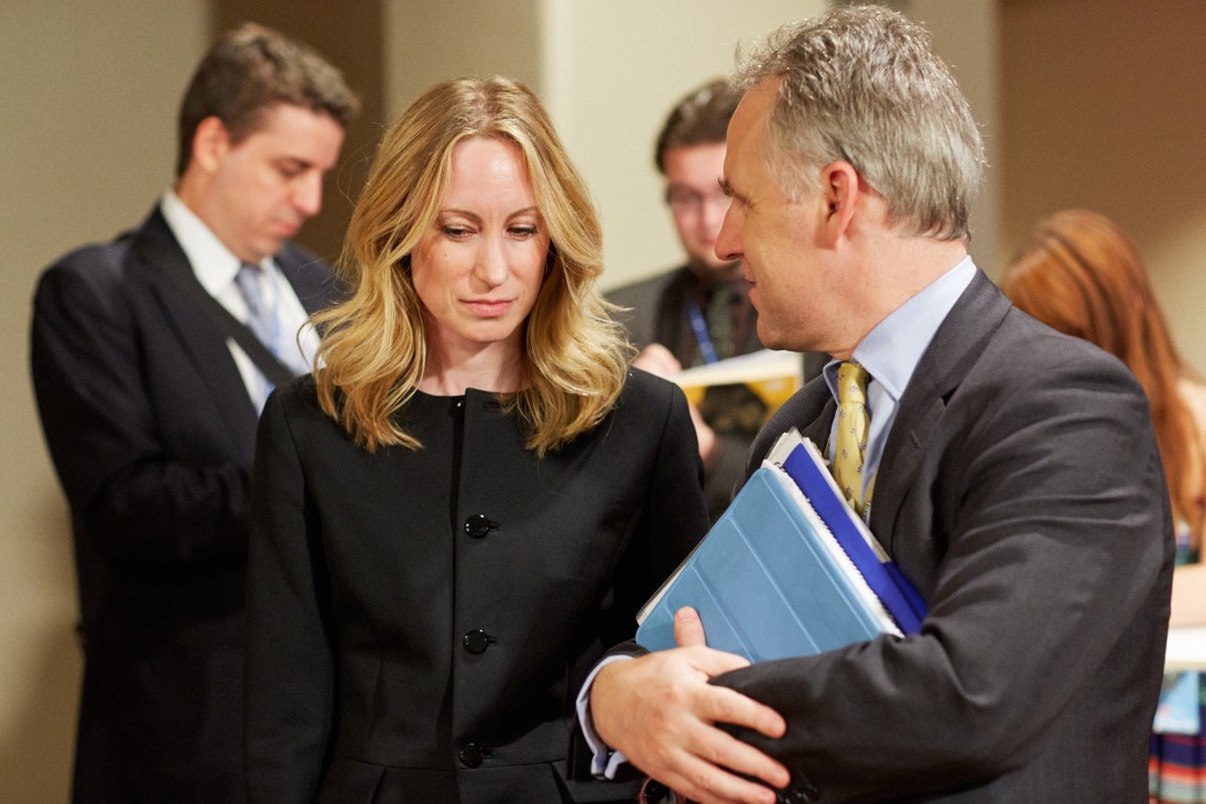 Two UK diplomats, Fiona Blyth and Michael Tatham, converse in a UN corridor while holding documents.