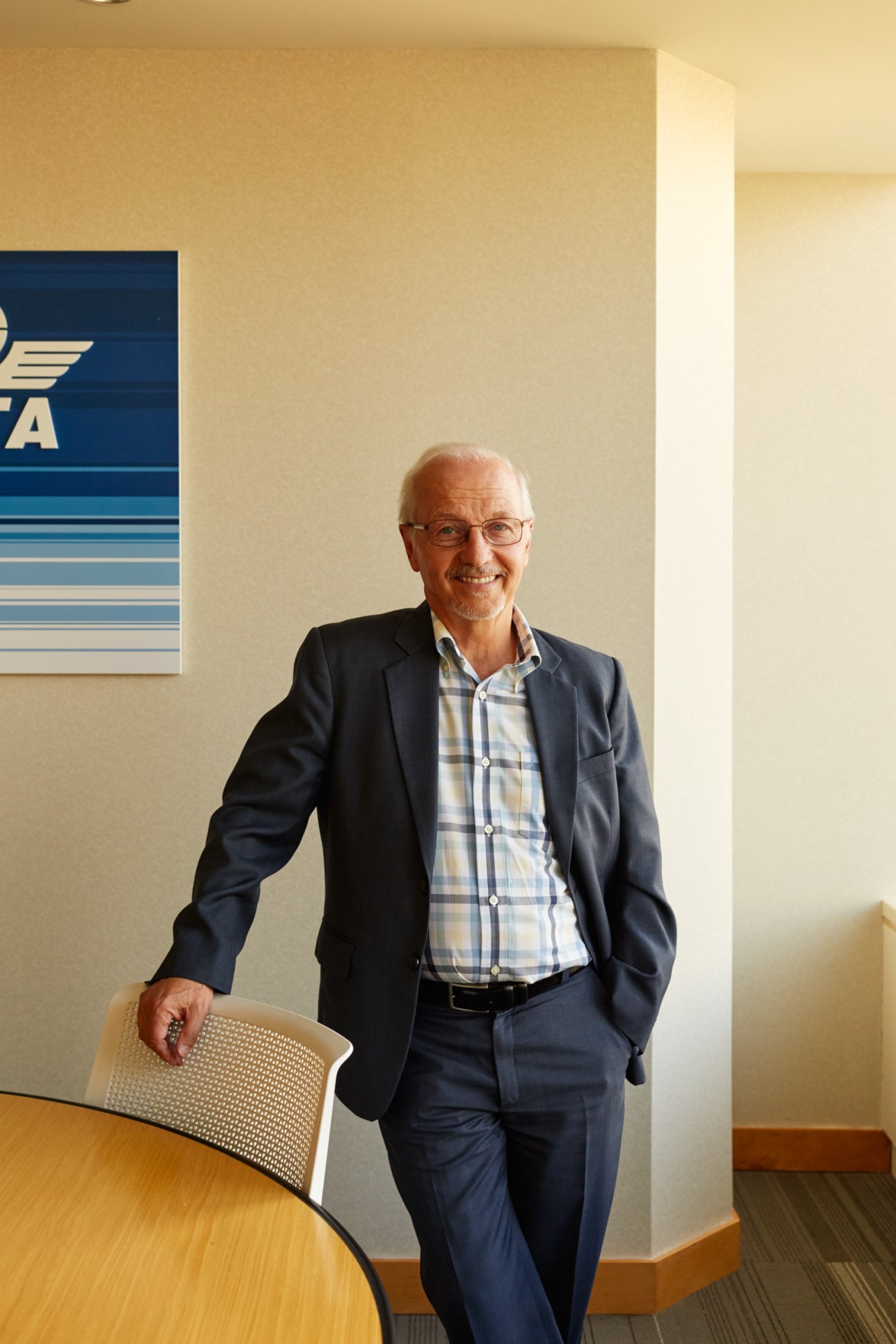 Guy Brazeau in navy blazer and checkered shirt standing beside office chair with IATA logo visible on wall behind him