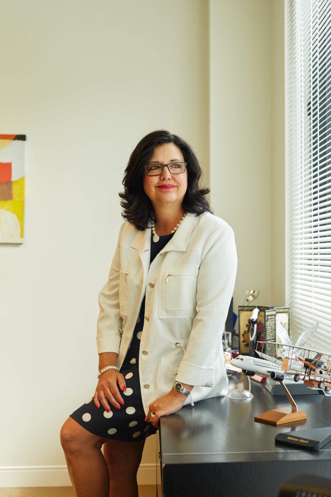 Suzanne Benoît sits at a desk in a modern office wearing a white blazer and glasses
