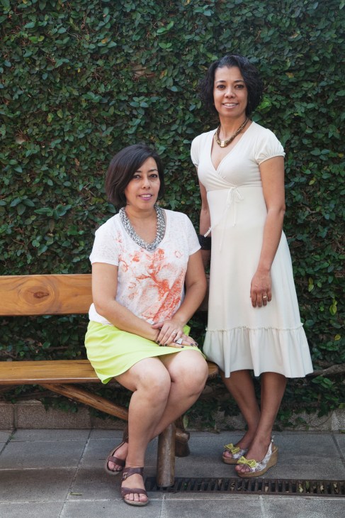 Two women smiling at camera in front of ivy-covered wall, one seated on wooden bench and one standing
