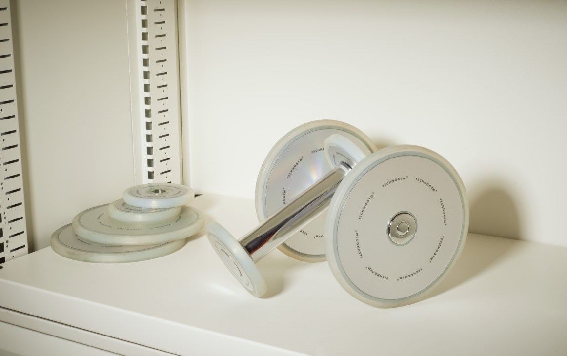 Technogym transparent dumbbells with silicone loadable disks displayed on a white shelf.