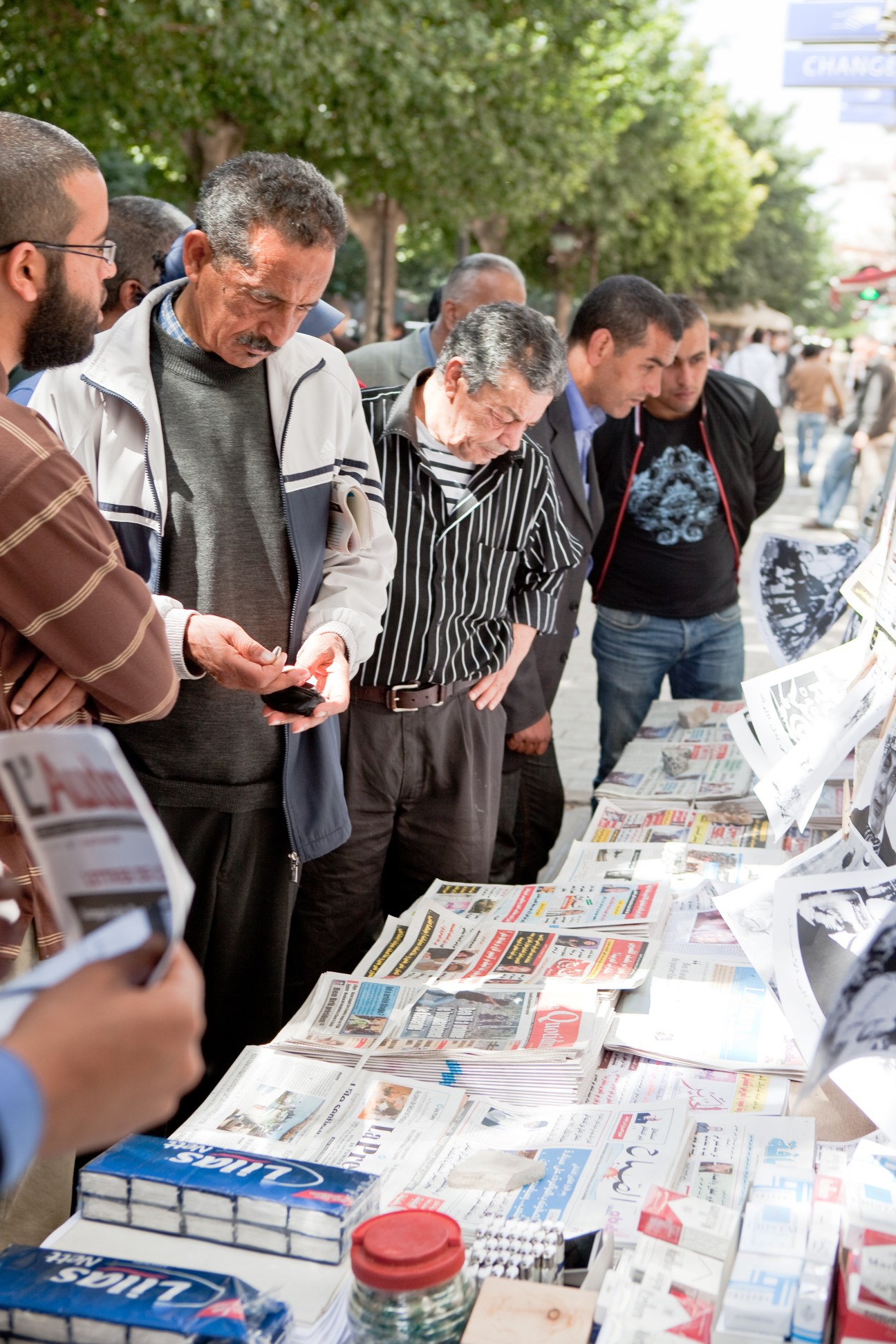 Men browsing newspapers displayed on a table at an outdoor market in Tunis.