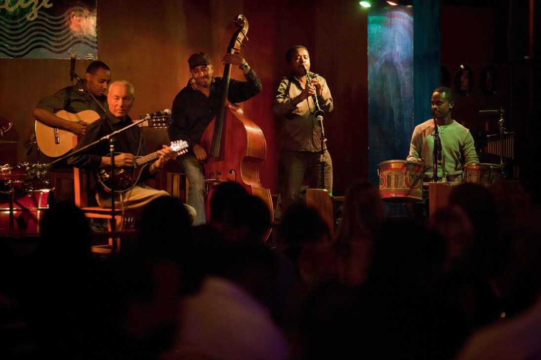 Five musicians performing on stage at Club Alizé with guitars, bass, and drums under warm stage lighting.