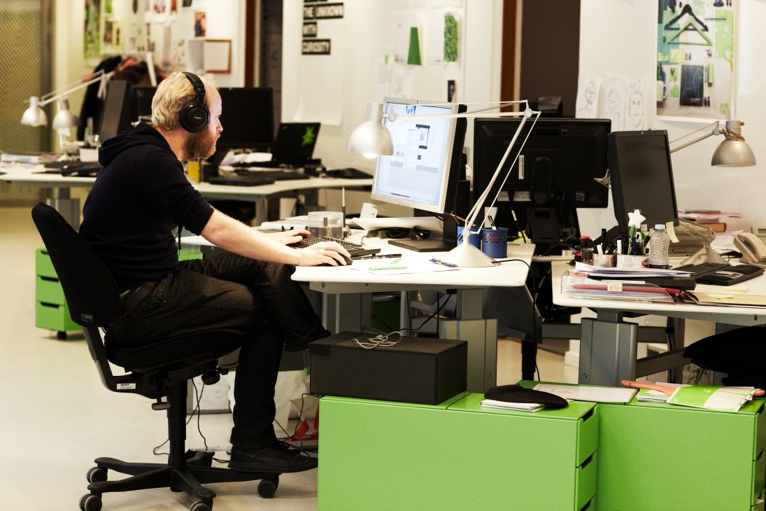 Man wearing headphones edits video at desk in modern office with green storage and design sketches on walls.