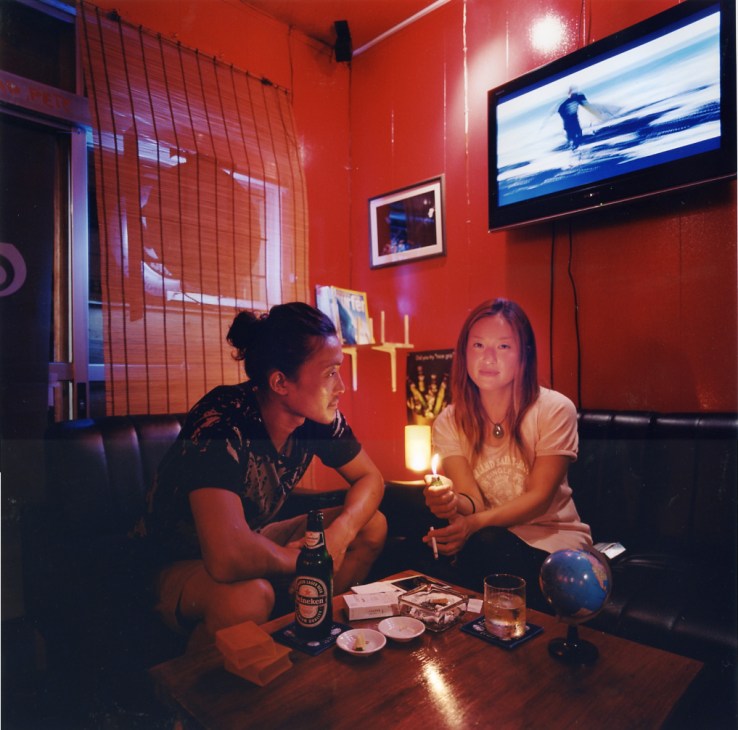 Two people sit in a red-walled bar with a TV showing a surfer, holding drinks and a glowing object.