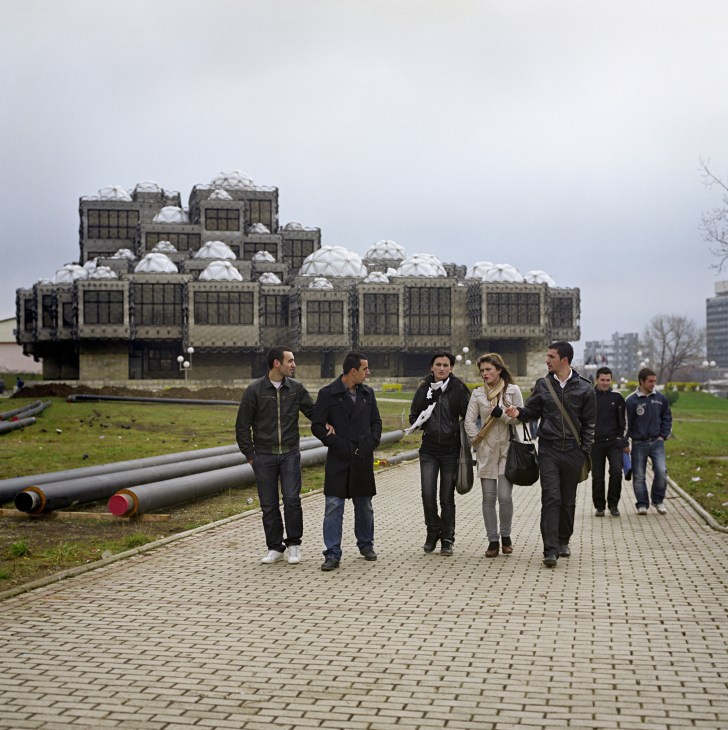 Students walking past Pristina's distinctive brutalist library building with white domes and geometric stone facade.