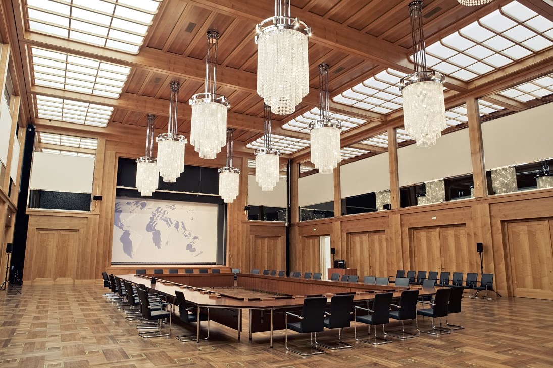 The Weltsaal conference room at Germany's Federal Foreign Office with wooden paneling, pendant chandeliers, and a large oval table.