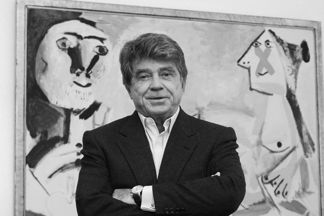 Man in dark suit and white shirt standing in front of framed artwork with stylized faces.