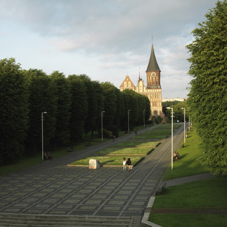 People walking through manicured green grounds toward a yellow neo-Gothic cathedral with a tall spire in Kaliningrad.